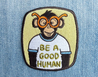 Puede incluir: Parche bordado para planchar con un mono de dibujos animados que lleva gafas de montura naranja y una camiseta blanca con el texto "Be a good human".
