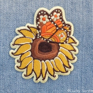 May include: Embroidered iron-on patch featuring a brown and orange butterfly with white spots, perched on a yellow sunflower with a brown center.