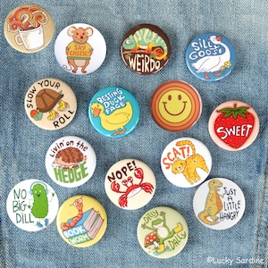 May include: A collection of fourteen circular pin-back buttons featuring various colorful cartoon illustrations and text, including a mouse saying Say Cheese, a turtle saying Slow Your Roll, a duck saying Resting Duck Face, and a dinosaur saying Just A Little Hangry.