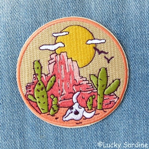 May include: Embroidered patch with a desert scene featuring a pink rock formation, a yellow sun, green cacti, and a white skull. The patch is circular and has a beige background.