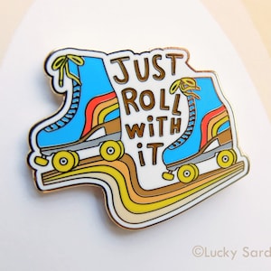 May include: Gold enamel pin featuring a pair of blue roller skates with rainbow stripes and wheels. The text "Just Roll With It" is written in a retro font.