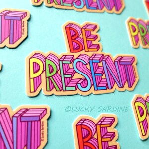 Peut inclure: Un ensemble d'autocollants color&eacute;s avec la phrase "Be Present" dans un style 3D. Les autocollants sont sur un fond bleu clair. Le texte est dans un sch&eacute;ma de couleurs arc-en-ciel avec du rose, du jaune, du vert et du bleu.