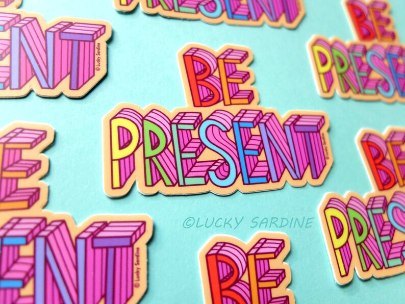 Peut inclure: Un ensemble d'autocollants color&eacute;s avec la phrase "Be Present" dans un style 3D. Les autocollants sont sur un fond bleu clair. Le texte est dans un sch&eacute;ma de couleurs arc-en-ciel avec du rose, du jaune, du vert et du bleu.
