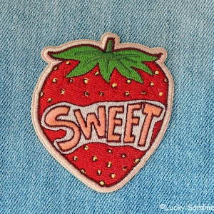 May include: A red and green embroidered iron-on patch shaped like a strawberry with the word "SWEET" in pink letters.