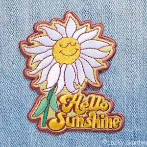 May include: Embroidered iron-on patch featuring a yellow daisy with a smiling face and the text "Hello Sunshine" in a retro font.