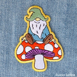 May include: Embroidered patch of a gnome sitting on a mushroom. The gnome is wearing a blue shirt and brown boots. The mushroom is red with white dots and has a purple cap.