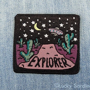 Peut inclure: Patch brodé noir avec une scène de désert rose, un croissant de lune, des étoiles, une soucoupe volante et deux cactus. Le texte "EXPLORER" est brodé en noir sur le patch.