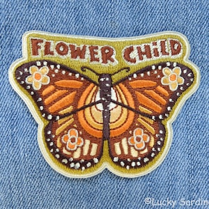 May include: Embroidered iron-on patch featuring a butterfly with orange, brown, and yellow wings. The butterfly has a white outline and is surrounded by a green border. The text "FLOWER CHILD" is written in brown letters on the top of the patch.
