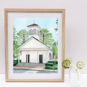 May include: A watercolor painting of a white church with a steeple and a bell tower. The church is surrounded by green trees and bushes. The painting is framed in a light wood frame.