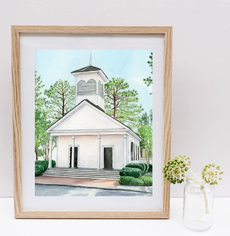May include: A watercolor painting of a white church with a steeple and a bell tower. The church is surrounded by green trees and bushes. The painting is framed in a light wood frame.