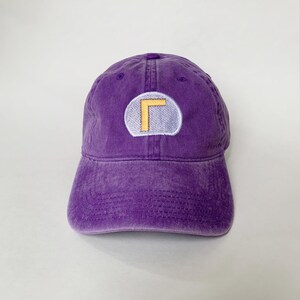K&ouml;nnte beinhalten: Eine lila Baseballkappe mit verwaschenem, vintage Aussehen. Die Kappe hat einen runden Aufn&auml;her mit gelbem und wei&szlig;em Design. Der Hut ist aus weichem Material und hat einen gebogenen Schirm.