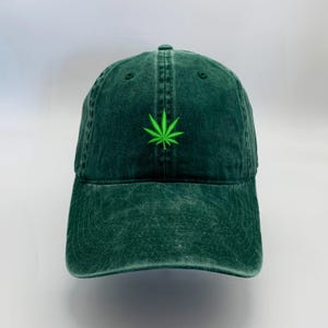 May include: A dark green, washed cotton baseball cap with a curved brim. The cap features a bright green embroidered cannabis leaf on the front. The hat has a classic six-panel design with an adjustable strap at the back.