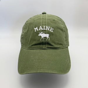 May include: A green baseball cap with white embroidery. The embroidery says "MAINE" and features a white moose silhouette.
