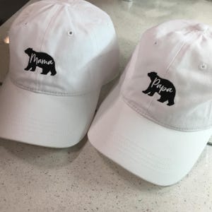 May include: Two white baseball caps featuring black embroidered bear silhouettes. One cap displays the word Mama in white script across the bear, while the other cap displays the word Papa in white script across the bear. Both caps have eyelets.