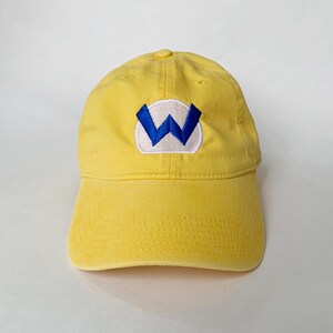 K&ouml;nnte beinhalten: Gelbe Baseballkappe mit gebogenem Schirm. Die Kappe hat einen wei&szlig;en Aufn&auml;her mit einem blauen "W" auf der Vorderseite. Die Kappe ist aus einem weichen, strapazierf&auml;higen Material gefertigt.