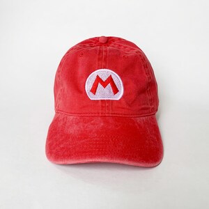 K&ouml;nnte beinhalten: Rote Baseballkappe mit einem wei&szlig;-roten gestickten "M"-Logo. Die Kappe hat einen gebogenen Schirm und einen stoffbezogenen Knopf. Der Hut ist aus weichem, abgenutztem Material.