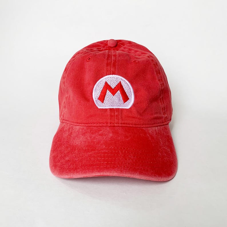 K&ouml;nnte beinhalten: Rote Baseballkappe mit einem wei&szlig;-roten gestickten "M"-Logo. Die Kappe hat einen gebogenen Schirm und einen stoffbezogenen Knopf. Der Hut ist aus weichem, abgenutztem Material.