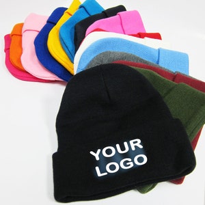 May include: A collection of knit beanies in various colors including black, white, gray, red, pink, orange, yellow, blue, and olive green. The front-most black beanie features the white text YOUR LOGO printed in a bold, sans-serif font on the cuff.