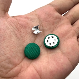 K&ouml;nnte beinhalten: Zwei gr&uuml;ne, stoffbezogene Kn&ouml;pfe mit einem silbernen Metall-Druckknopf. Die Kn&ouml;pfe sind rund und haben eine strukturierte Oberfl&auml;che.