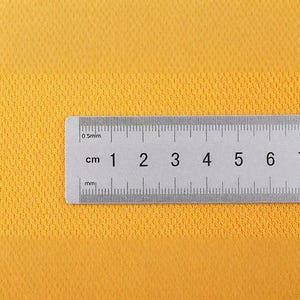 May include: A close-up shot of a yellow textured fabric with a ruler placed horizontally across the bottom. The ruler is marked in centimeters, with numbers from 1 to 7 visible. The fabric has a subtle, grid-like pattern.