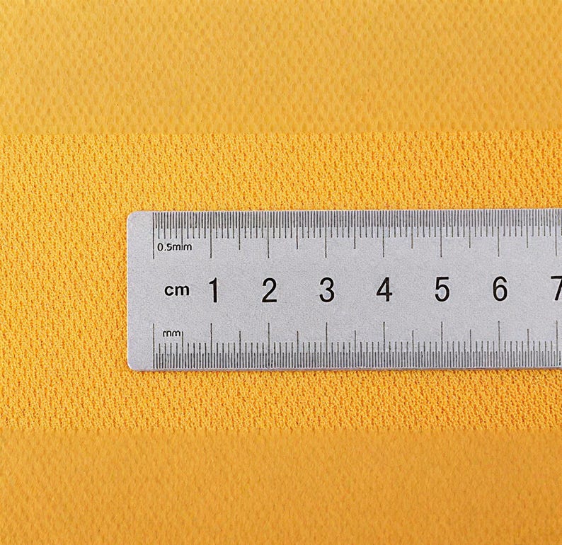 May include: A close-up shot of a yellow textured fabric with a ruler placed horizontally across the bottom. The ruler is marked in centimeters, with numbers from 1 to 7 visible. The fabric has a subtle, grid-like pattern.