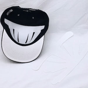May include: A black and white baseball cap with a white brim and a black crown. The crown has a white design that resembles a smile. The cap is lying on a white surface next to a set of white paper cutouts.