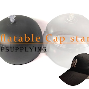 Puede incluir: Dos soportes de gorra inflables, uno negro y otro transparente, con una gorra de béisbol negra con un logotipo blanco en la parte superior del soporte transparente. El texto "Inflatable Cap stand" y "CAPSUPPLYING" es visible en la imagen.