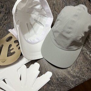May include: Two baseball caps, one white and one gray. The white cap has a white brim and a white strap. The gray cap has a gray brim and a gray strap. The gray cap has the text "ADIDAS" on the strap.