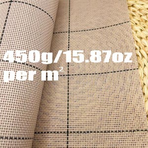 May include: Close-up of a beige rug canvas with a grid pattern. The text "450g/15.87oz per m²" is printed on the canvas. The rug canvas is partially rolled, revealing its texture and construction.