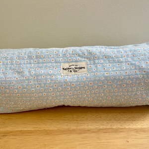 May include: A rectangular, light blue fabric pouch with a floral pattern of small white and orange dots. The pouch has a rectangular label that reads "Made by Kaylee's Designs & Co." and two small fabric handles on each end.
