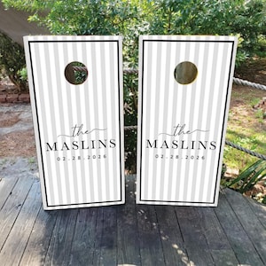 May include: Two rectangular cornhole boards featuring a vertical light gray and white striped pattern with a black border. Each board displays the text the MASLINS and the date 02.28.2026 in black lettering, centered below a circular hole.