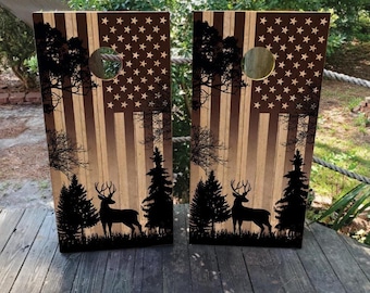 May include: Two rectangular cornhole boards featuring a wood grain finish with a dark brown American flag design. Each board displays a black silhouette of a deer standing in a forest with pine trees and a circular hole near the top center.