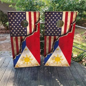May include: Two wooden cornhole boards. Each board features a design that combines the American and Philippine flags. The top half displays the American flag, while the bottom half shows the Philippine flag. Each board has a circular hole.