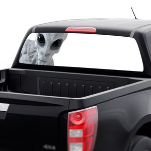 May include: A black pickup truck with a rear window decal of a gray alien peeking out from behind the window.