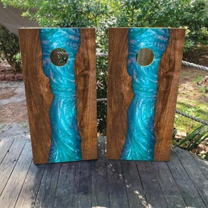 Könnte beinhalten: Zwei rechteckige Cornhole-Boards mit natürlichen braunen Holzkanten und einem zentralen Flussdesign aus wirbelndem, durchscheinendem türkisfarbenem und blaugrünem Harz. Jedes Board hat ein kreisförmiges Loch im oberen Bereich.