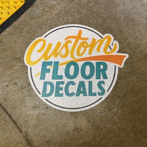 May include: Circular floor decal with the words "Custom Floor Decals" in a stylized font. The word "Custom" is in yellow and orange, while "Floor Decals" is in teal. The decal has a white background and a black border.