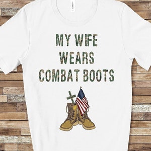 May include: White t-shirt with the text "My Wife Wears Combat Boots" in a camouflage font. A pair of brown combat boots with an American flag are shown below the text.