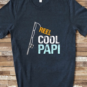 May include: A dark blue t-shirt with a white fishing rod graphic and the text "REEL COOL PAPI" in orange and blue.