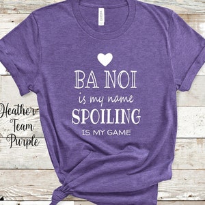 May include: Purple t-shirt with white text that reads "Ba Noi is my name Spoiling is my game". A white heart is above the text.