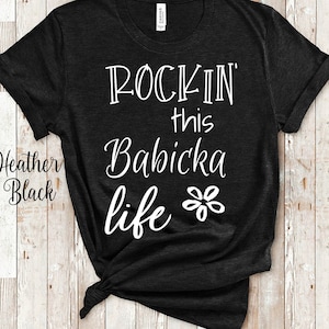 Puede incluir: Camiseta negra de color jaspeado con texto blanco que dice "Rockin' this Babicka life" y un gráfico de flor blanca.