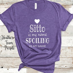 May include: Heather Team Purple t-shirt with the text "Sitto is my name, spoiling is my game" in white. A white heart is above the word "Sitto". The shirt is knotted at the bottom.