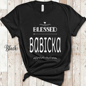 Puede incluir: Camiseta negra con la frase "BLESSED BABICKA" en blanco, con elementos decorativos. La camiseta está hecha de un material suave y es adecuada para ropa casual. La palabra "Black" está escrita en el lateral de la camiseta.