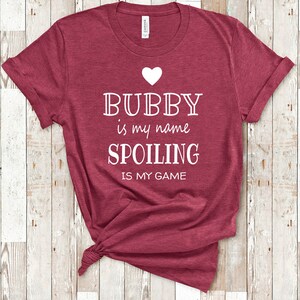 May include: Burgundy red t-shirt with white text that reads "Bubby is my name Spoiling is my game" with a small white heart above the text.