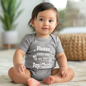 May include: A gray baby onesie with the text "Please pass me to my Papi Chulo" in white. The onesie has short sleeves and is decorated with small heart shapes. The baby is sitting on a light-colored rug.
