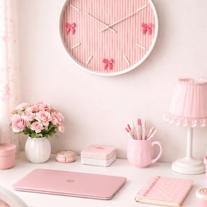 May include: A pink and white striped wall clock with a white frame and pink bow accents. The clock is part of a desk setup that includes a pink laptop, a pink mug, a notepad, and a lamp.