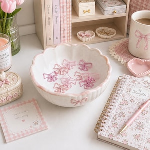 May include: Aesthetic desk scene with a white scalloped bowl holding pink bow-shaped paper clips. A floral notebook, a pink pen, and a mug with a bow design are also present. A candle and a small trinket box add to the feminine theme.