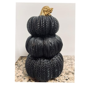 Könnte beinhalten: Ein dekorativer schwarzer Kürbisstapel mit goldenem Stiel. Die Kürbisse haben ein strukturiertes, gewebtes Aussehen. Der Stapel besteht aus drei unterschiedlich großen Kürbissen und schafft eine einzigartige herbstliche Dekoration.