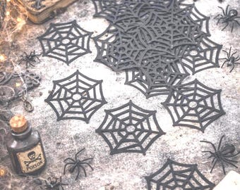 May include: A Halloween-themed arrangement featuring black spiderweb cutouts, plastic spiders, a skull, and a vintage book with the word "SPELLS". A candle burns beside the book, and a small bottle labeled "POISON" is also present.