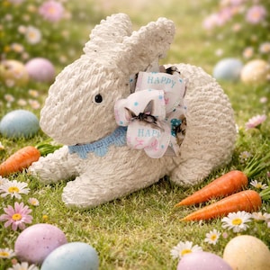 May include: A white Easter bunny figurine with a blue collar and a bow that says "HAPPY". The bunny is surrounded by Easter eggs, carrots, and flowers, creating a festive spring scene.