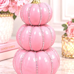 May include: A stack of three glossy light pink pumpkins with a metallic gold stem. Each pumpkin is decorated with vertical rows of clear, sparkling rhinestones along the ridges. The pumpkins are arranged in a vertical tower formation.